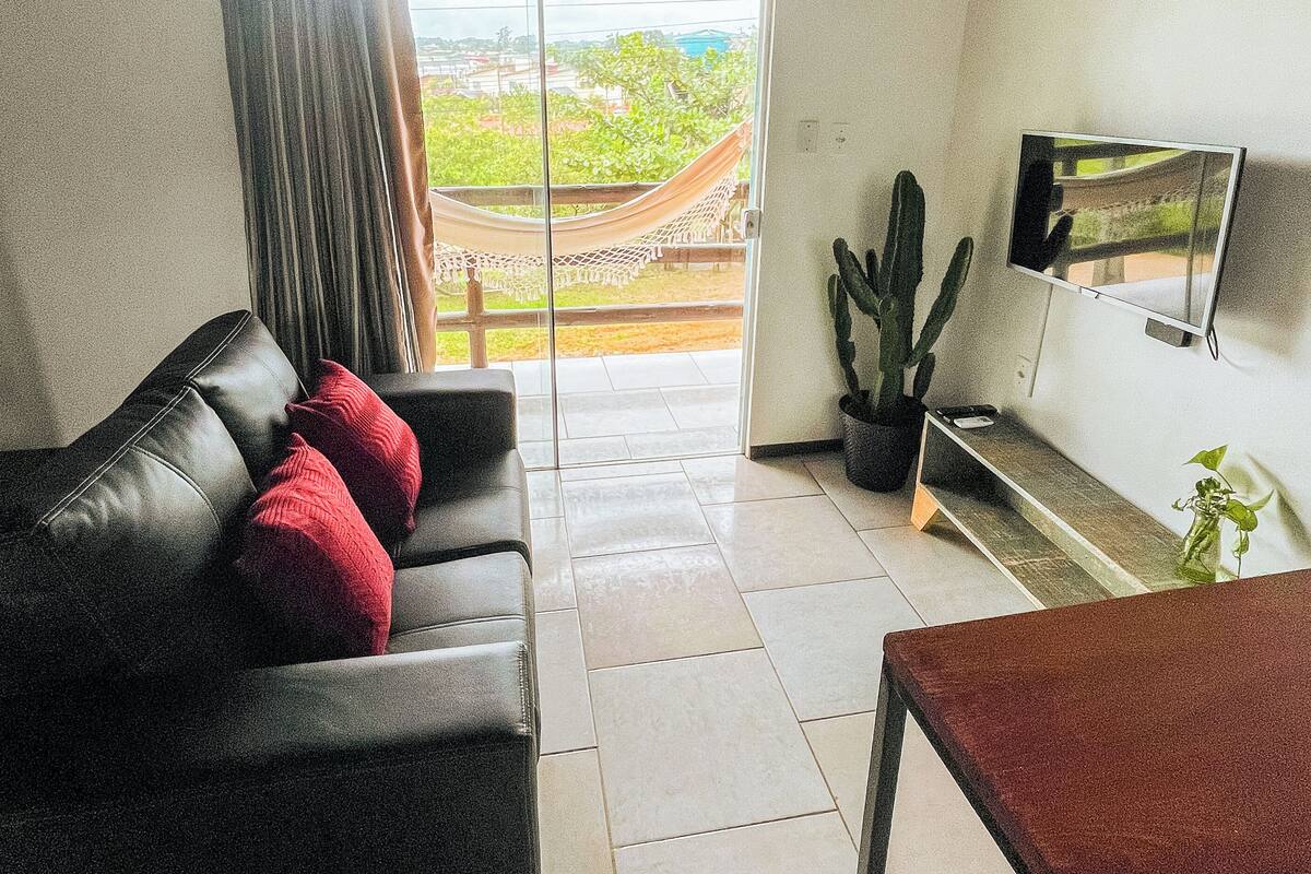 A living room with a black leather sofa, red cushions, a wooden coffee table, a TV mounted on the wall, and a view of greenery through a glass door.