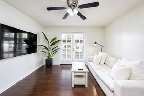 Una sala de estar moderna con un sofá blanco, una mesa de centro, una planta en maceta, un ventilador de techo y un ventanal con persianas.