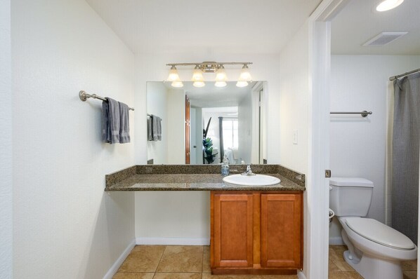 A bathroom with a granite countertop, wooden cabinets, a white sink, a mirror with lights, and a toilet.