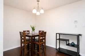 Un área de comedor con una mesa de madera, cuatro sillas, una planta en maceta, un estante de metal negro y una lámpara en el techo.