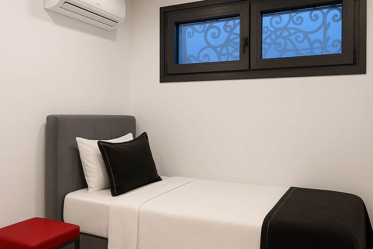 A modern bedroom with a grey bed, white bedding, a black throw, a red stool, and an air conditioning unit on the wall.