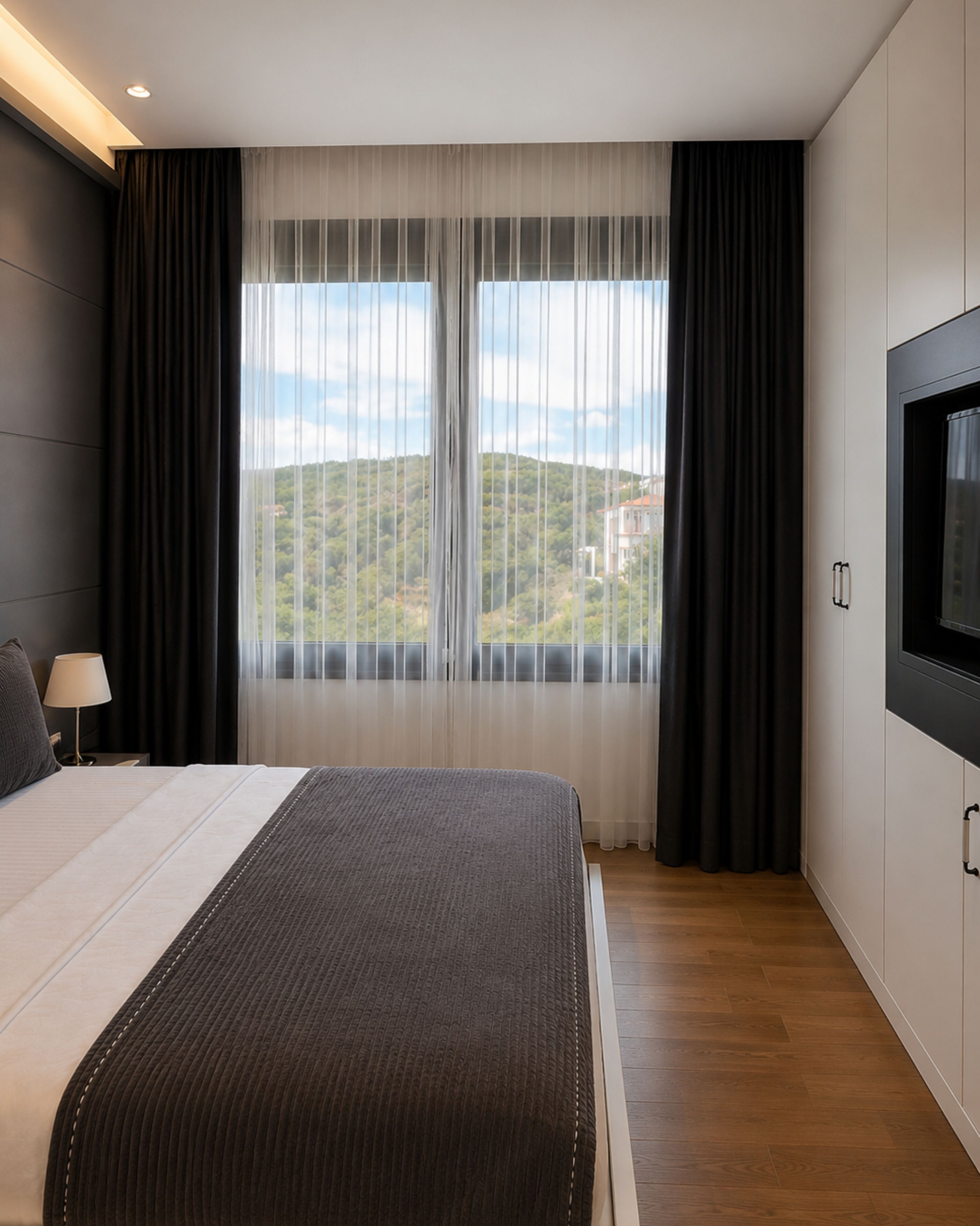 A hotel room with a large bed, a TV, and a view of a hilly landscape through sheer curtains.