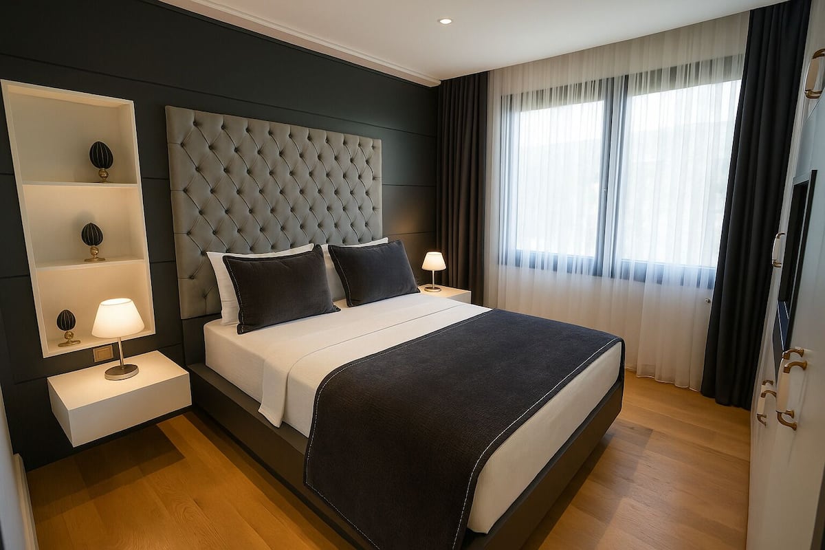 A modern hotel room with a tufted headboard, a large bed with a black throw, two bedside tables with lamps, and a large window with sheer curtains.