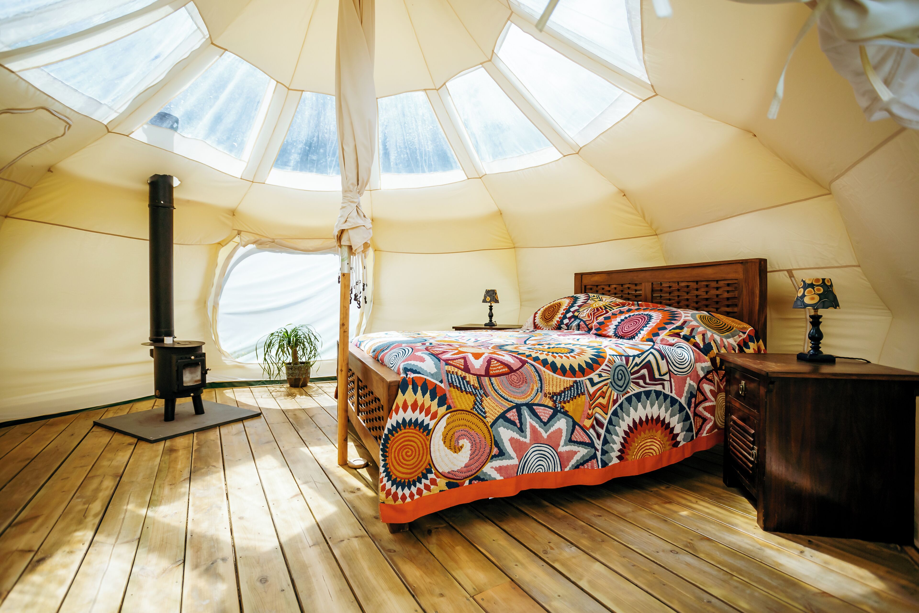 A cozy tent-style bedroom with a wooden floor, a colorful bedspread, a small table, a lamp, and a potted plant.