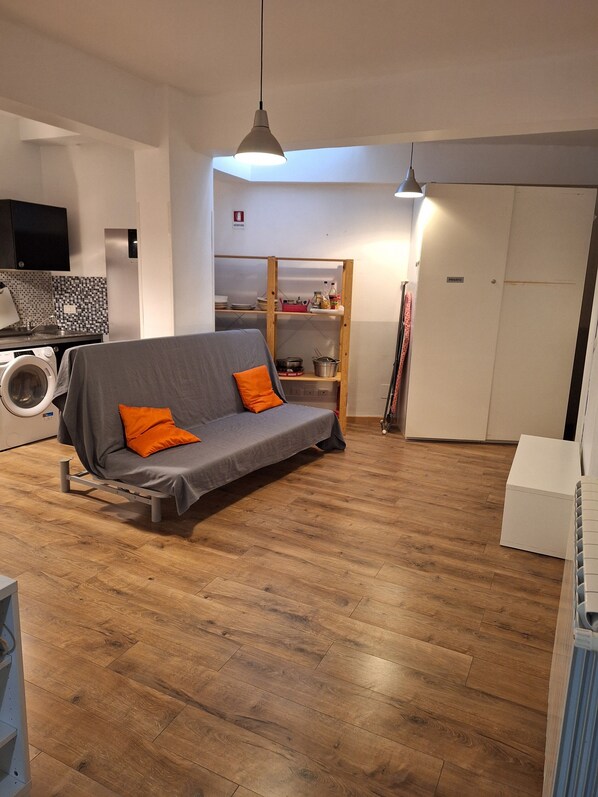 A modern living space with a grey sofa, wooden flooring, a washing machine, and a built-in shelf with kitchen items.