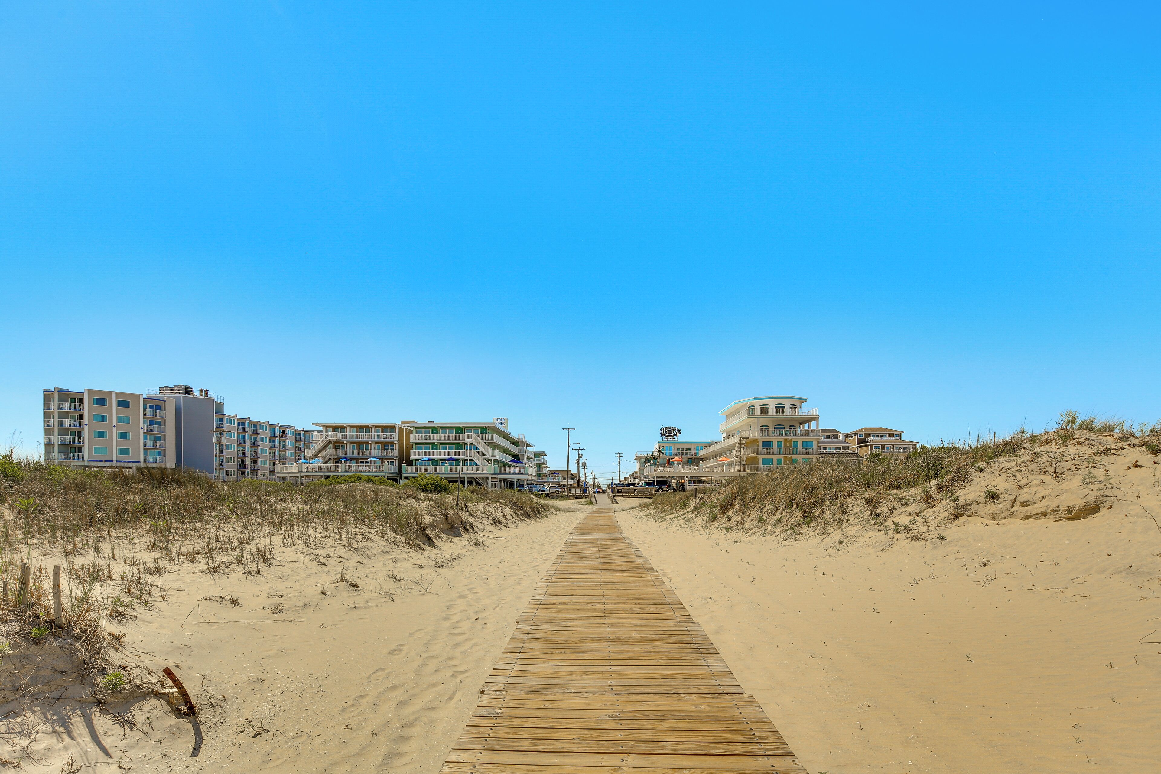 Ein hölzerner Steg führt zu Strandhäusern mit mehreren Stockwerken und Balkonen.