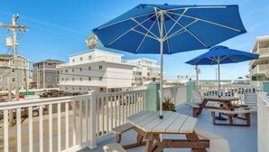 Eine Terrasse am Meer mit Holztischen und blauen Sonnenschirmen, mit Blick auf eine Straße mit Gebäuden und blauem Himmel.