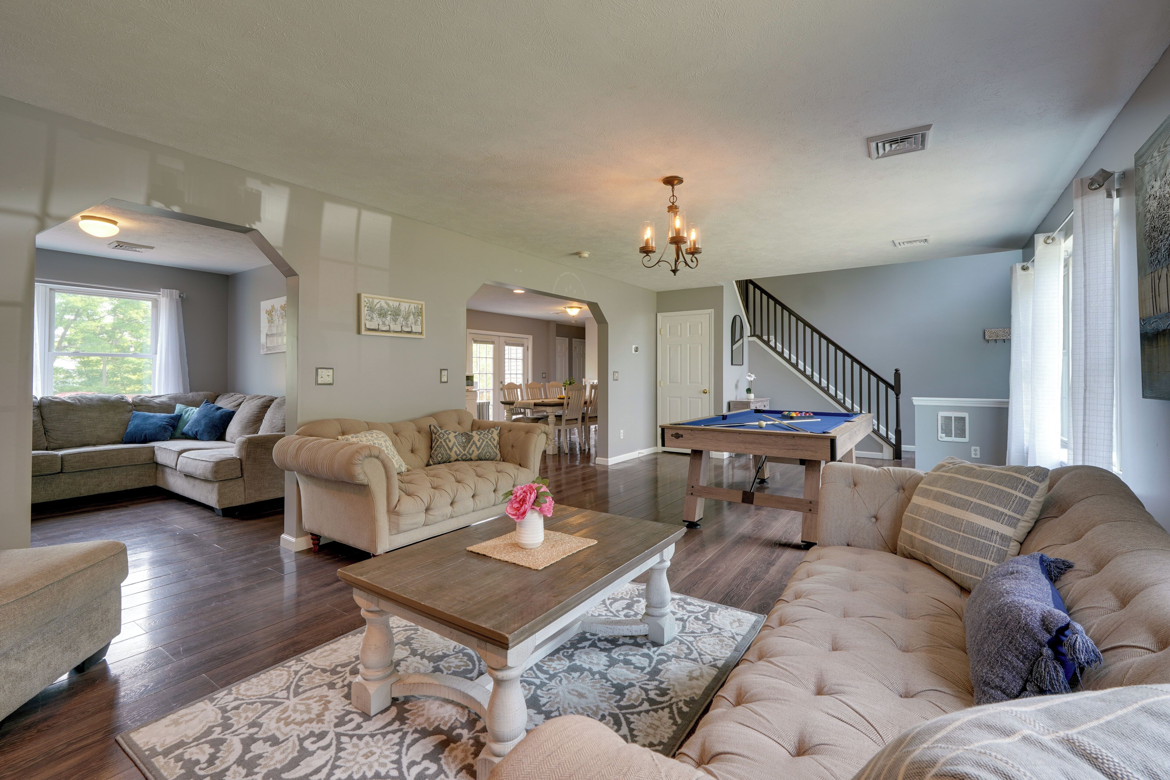 A spacious living room with a large sofa, a coffee table, and a pool table. There is a staircase leading to another level.