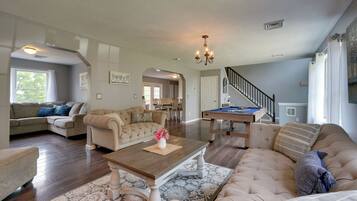 A spacious living room with a large sofa, a coffee table, and a pool table. There is a staircase leading to another level.