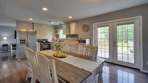 Une cuisine spacieuse avec une table à manger en bois, des chaises blanches et un panier de fruits. La cuisine est équipée d’un réfrigérateur, d’un four à micro-ondes et d’un four traditionnel.