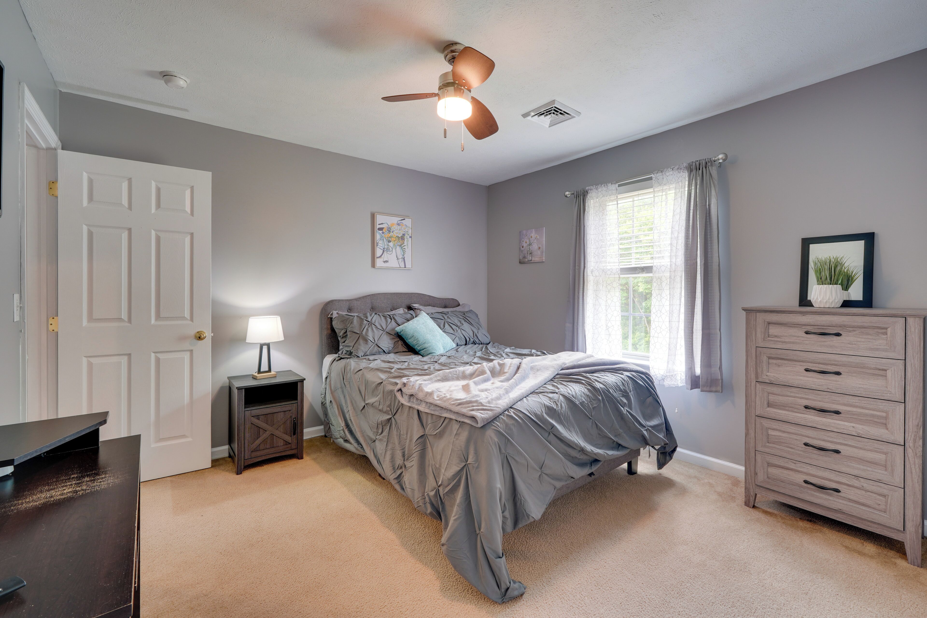 A bedroom with a bed, a dresser, a nightstand, a ceiling fan, and a window with curtains.