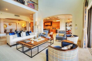 A spacious living area with a white sofa, blue armchairs, a wooden coffee table, and a kitchen in the background.