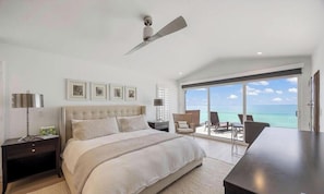 A bedroom with a large bed, a ceiling fan, a nightstand, a desk, and a view of the ocean.