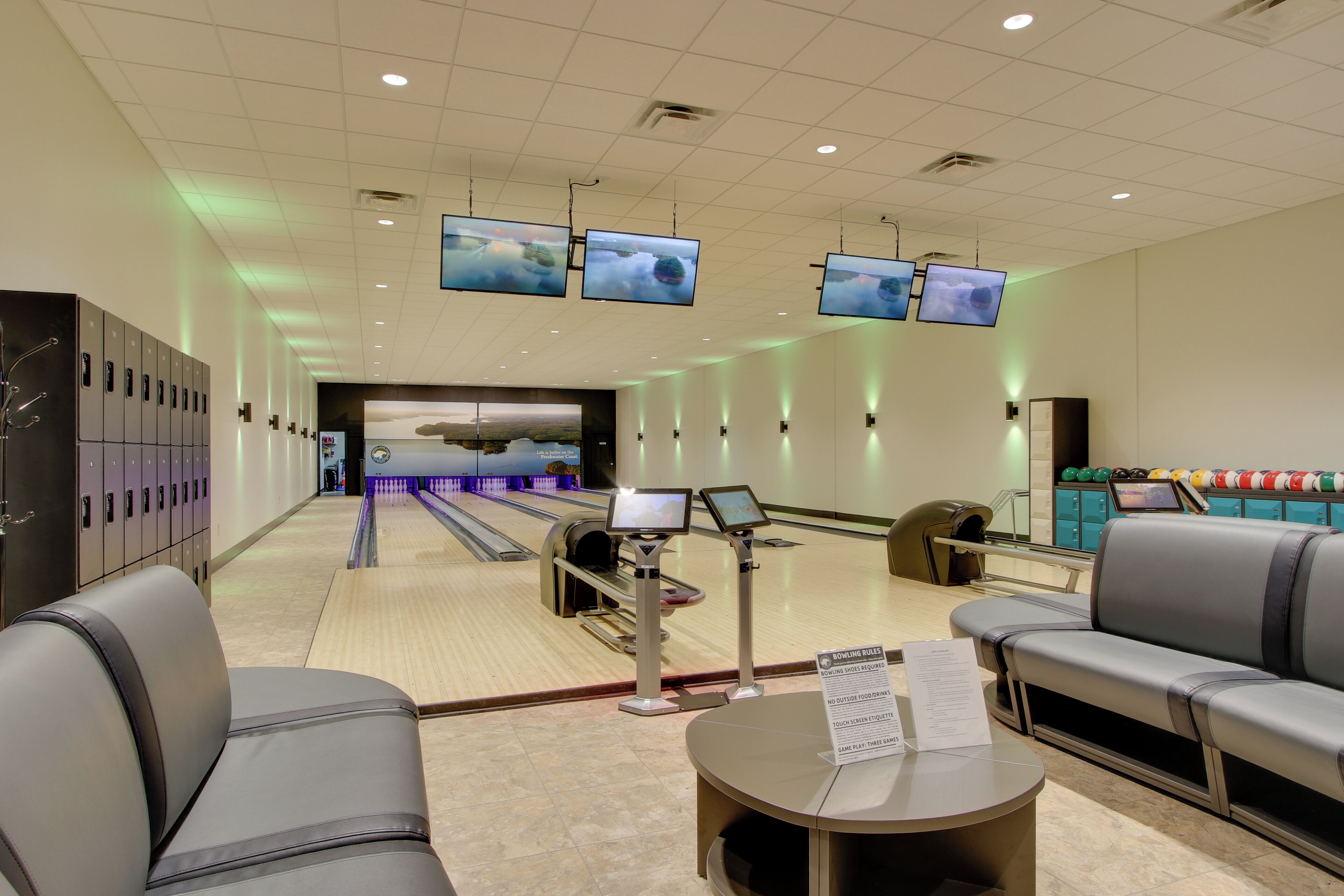 Una pista da bowling con numerose piste, un'area salotto con divani grigi e diversi monitor che mostrano immagini.