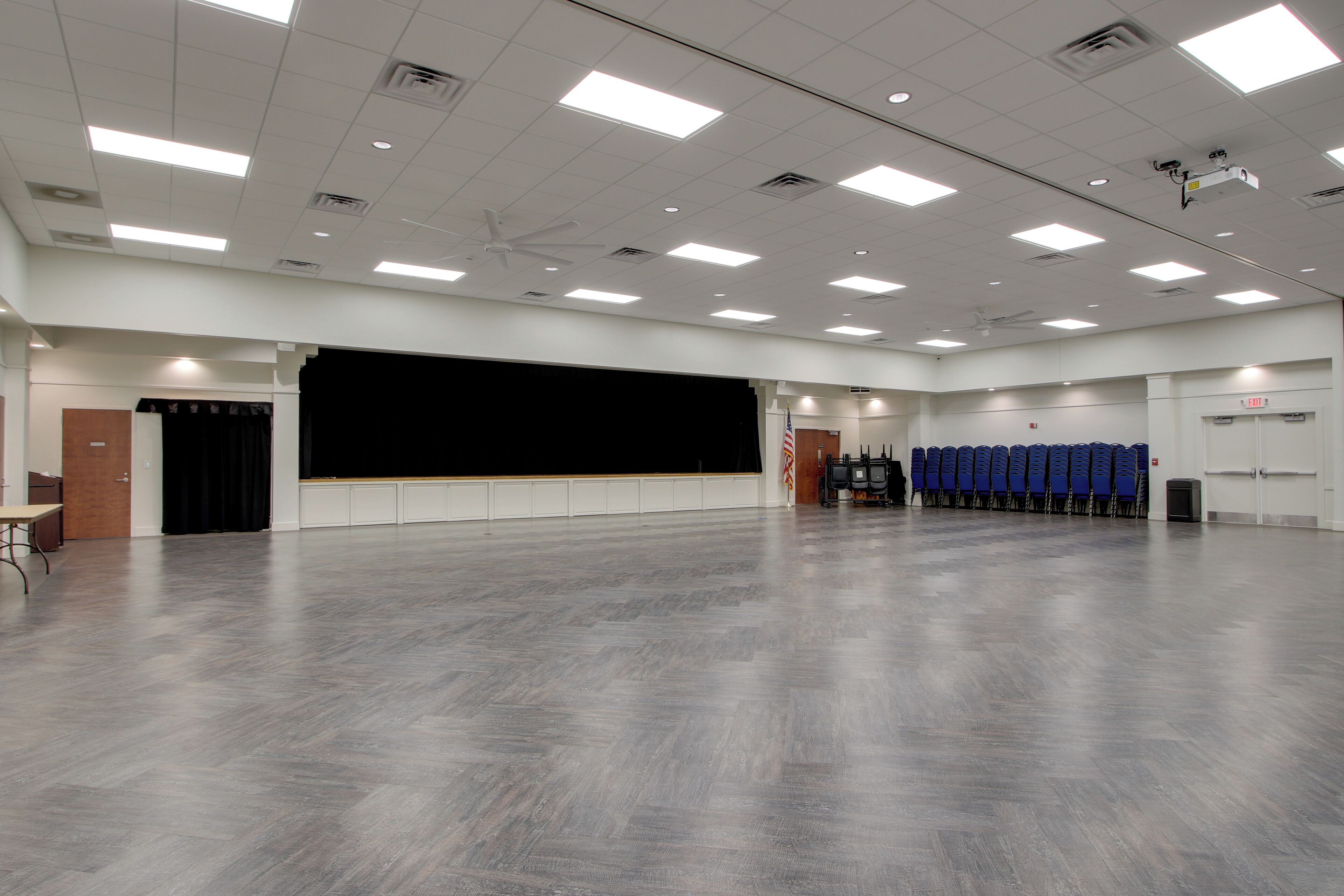 A spacious room with a stage, ceiling fans, and rows of blue chairs.