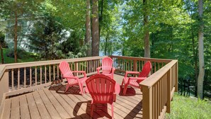 Een houten terras met rode Adirondack-stoelen en uitzicht op een meer, omgeven door bomen.
