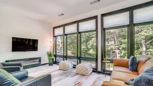 A modern living room with a flat-screen TV, leather sofas, and large windows offering a view of trees. - Game Room, Fire Pit Free Pickleball & Tennis (Helen)