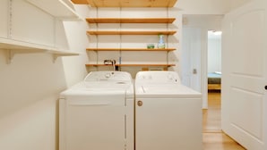 Un cuarto de lavandería con dos lavadoras blancas, estantes de madera arriba y una puerta que da a otra habitación.