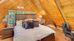 Ein gemütliches Schlafzimmer mit holzverkleideter Schräge, einem großen Bett mit gemusterten Kissen, einem Nachttisch mit Lampe und einem Sessel.