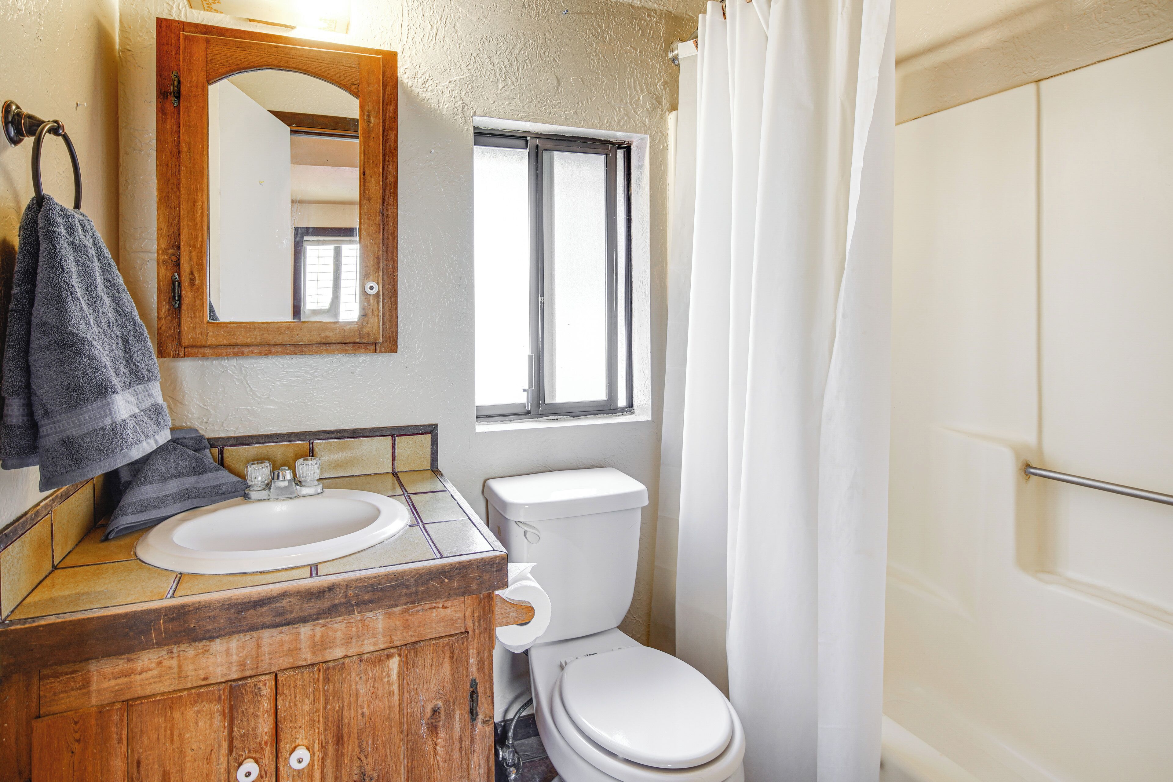 Ein Badezimmer mit einem Holzbadezimmerhocker, einem weißen Waschbecken, einer Toilette und einem Fenster mit Blick nach draußen.
