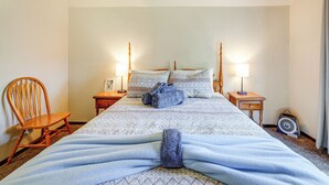 Una cama bien hecha con una colcha estampada y almohadas, una silla de madera, mesitas de noche con lámparas y un ventilador en el suelo.