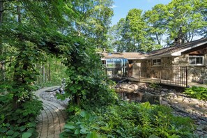 A stone house with a glass conservatory, surrounded by lush greenery and a stone pathway.