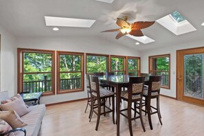 A dining area with a table and chairs, large windows with a view of trees, a ceiling fan, and a door with a glass panel.