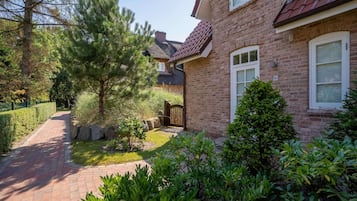 Une maison en briques avec un chemin pavé, un petit jardin agrémenté de différentes plantes et un portail en bois.