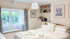 Ein Schlafzimmer mit einem großen Bett, einem Nachttisch, einem Fenster mit Blick auf Grünflächen, einem Bücherregal und einem gerahmten Bild an der Wand.