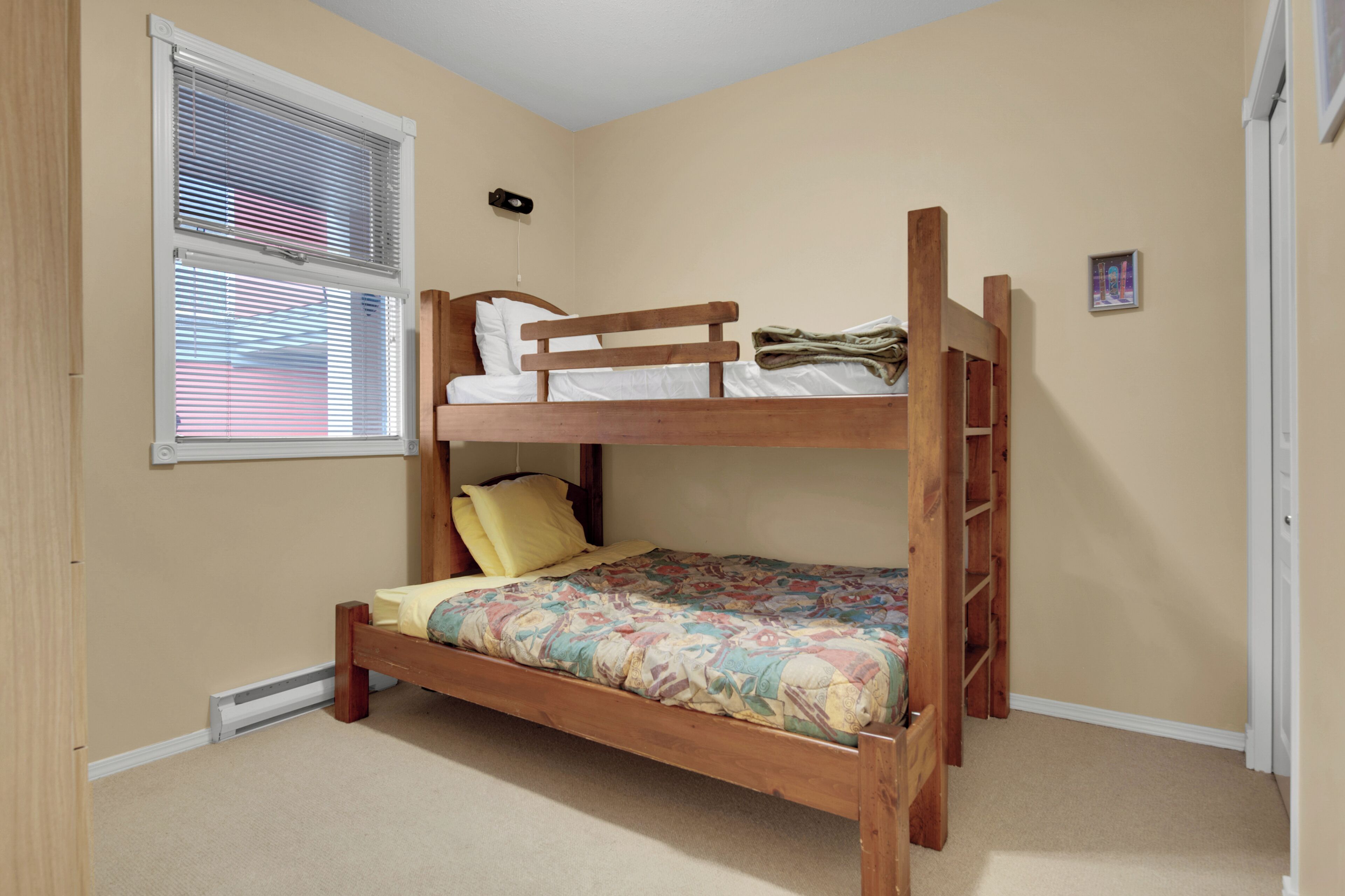 A bunk bed in a room with beige walls, a window with blinds, and a door.