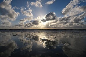 A beach with a reflective wet surface, scattered clouds, and the sun shining through.