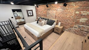 A bedroom with a brick wall, a wooden bed, a nightstand, and a staircase. - DTSF Historic Phillips Loft in the heart of Downtown Sioux Falls (Sioux Falls)