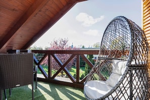 Un espacio exterior techado con un sillón de mimbre y una mesa, con vistas a un jardín con árboles y un cielo despejado.