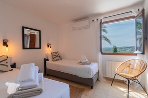 Een hotelkamer met twee bedden, een groot raam met uitzicht, een rieten stoel en een spiegel aan de muur.