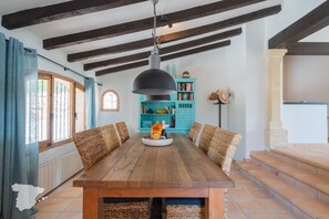 A wooden dining table with woven chairs, a hanging pendant light, and a turquoise bookshelf in a room with exposed wooden beams and a tiled floor.