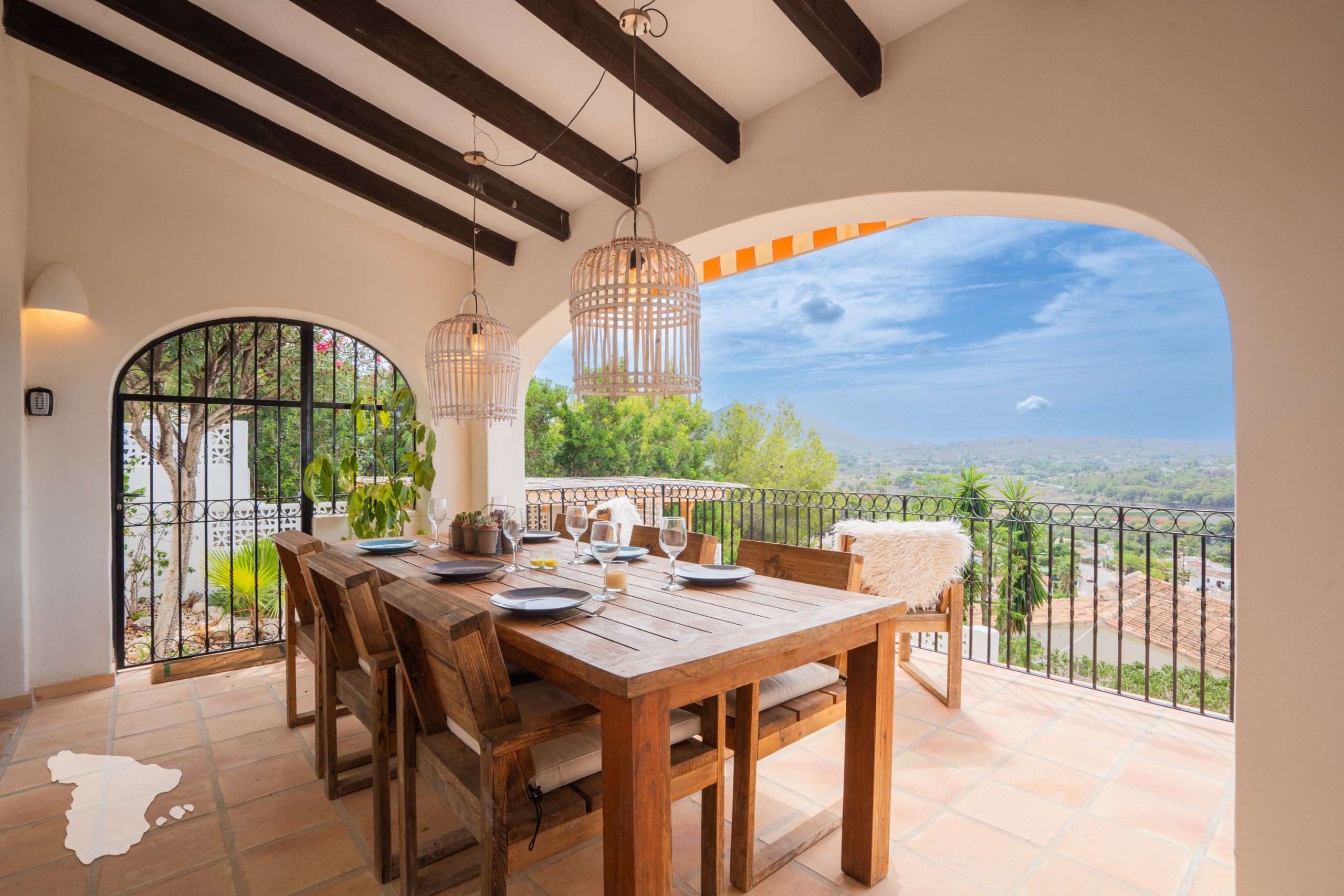 A wooden dining table set with plates, glasses, and a candle, overlooking a scenic view with a balcony and railing.