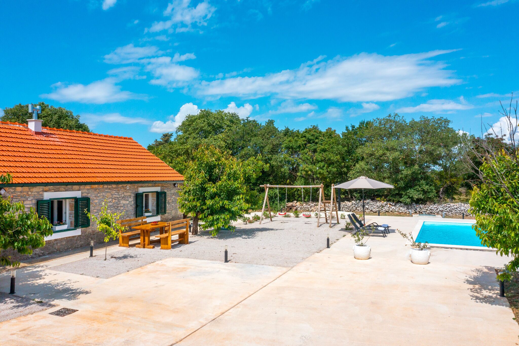 A stone house with a red roof, a swimming pool, a wooden bench, and a swing set.