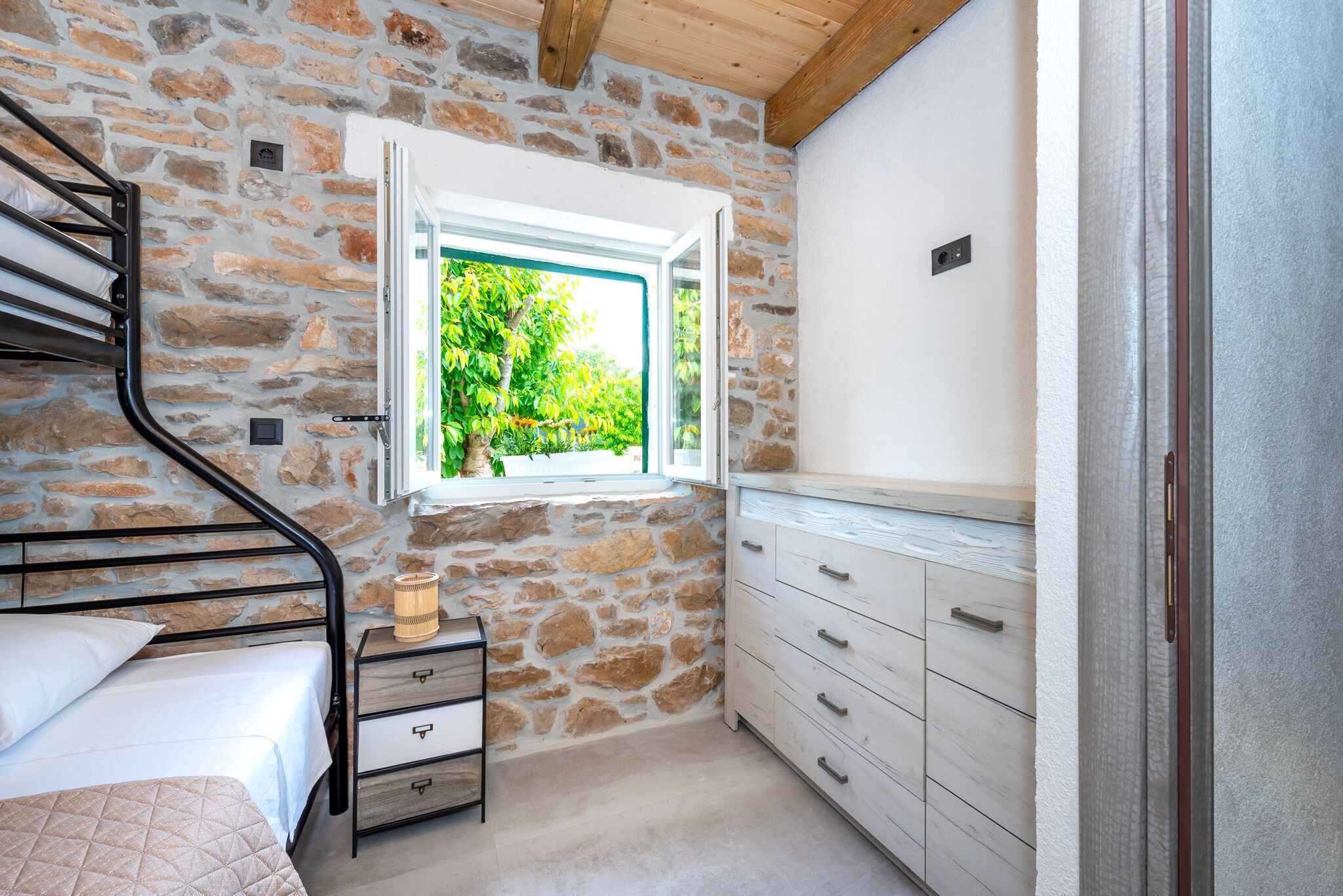 A bedroom with a stone wall, a bed with a headboard, a nightstand, a dresser, and a window with a view of greenery.
