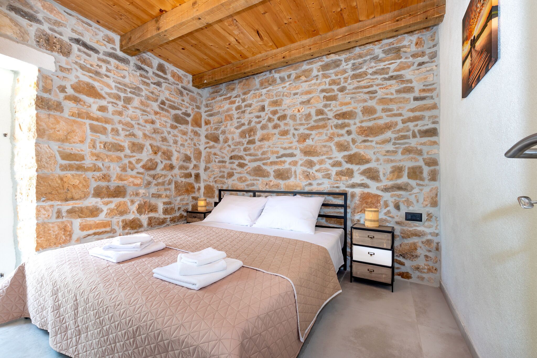A bedroom with a stone wall, wooden ceiling, a bed with a beige quilt, white pillows, and a nightstand with a lamp.