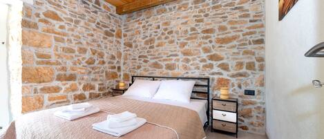 A bedroom with a stone wall, wooden ceiling, a bed with a beige quilt, white pillows, and a nightstand with a lamp.