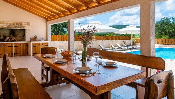 A wooden dining table set for four with a stone fireplace, wooden cabinets, and a view of a swimming pool and mountains. - Villa Kole (Konjevrate)