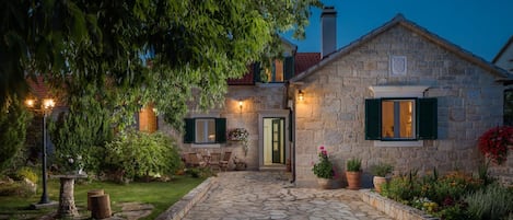 A stone house with a stone pathway leading to the entrance, surrounded by greenery and potted plants.