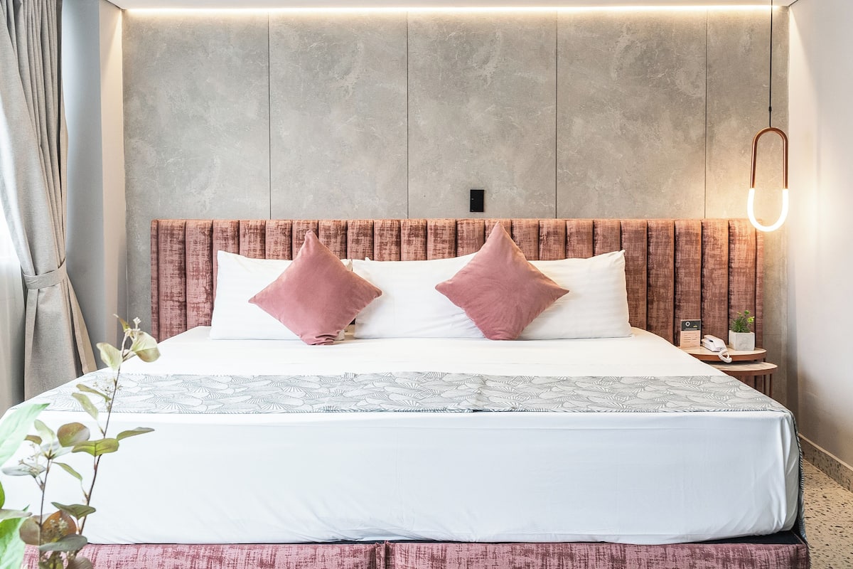 Quarto de hotel moderno com uma cama grande, cabeceira de madeira e mesa de cabeceira com telefone e planta.