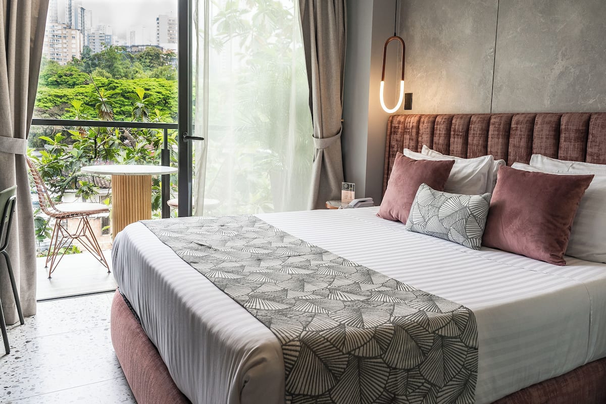 Quarto de hotel moderno com uma cama grande, varanda com vista para áreas verdes e edifícios, e uma pequena mesa.