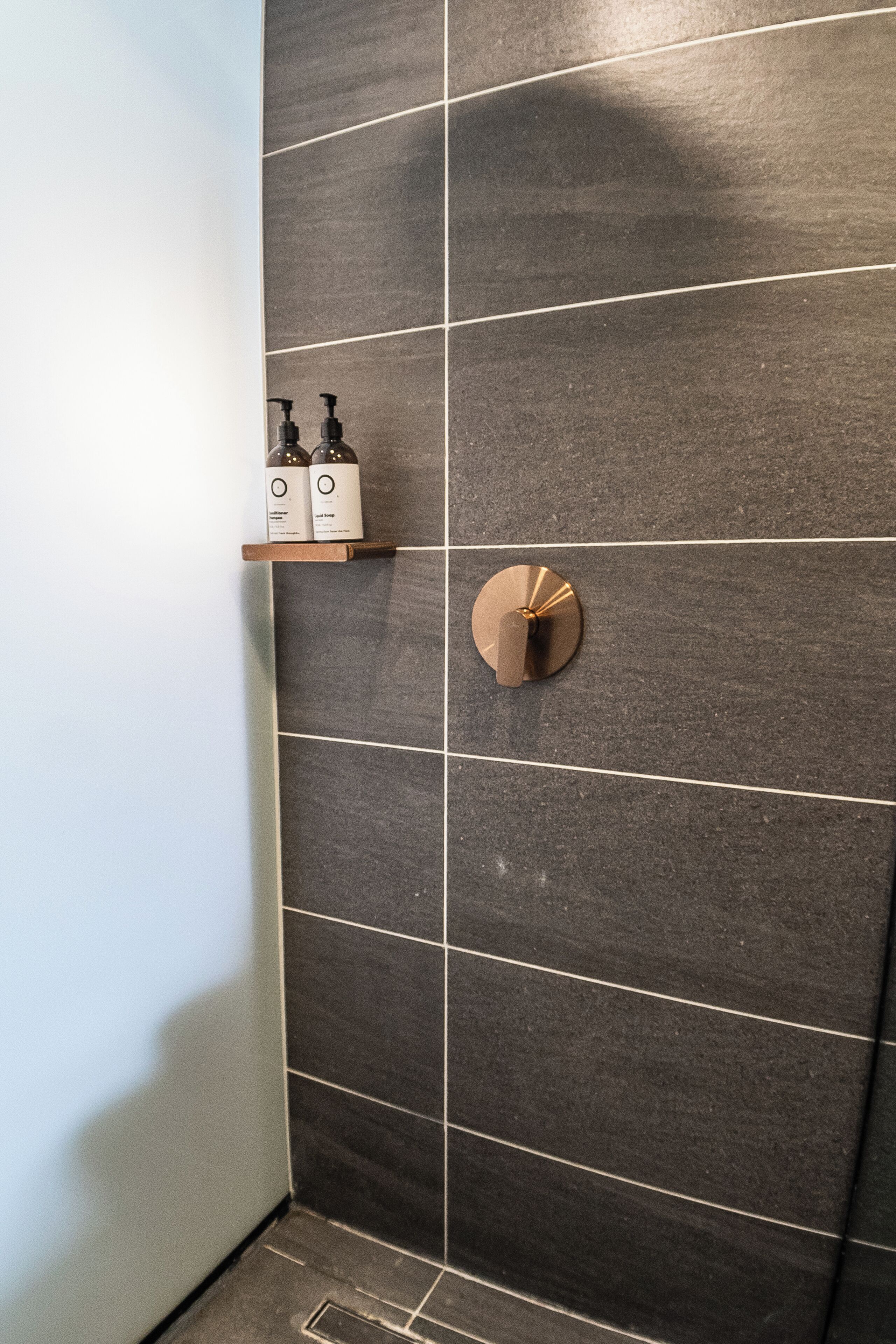 Un baño con paredes de azulejos oscuros, un estante de madera con artículos de aseo y un objeto redondo metálico en la pared.