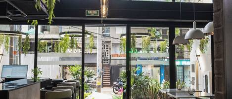 A modern cafe with a view of a building across the street, featuring a reception area, seating, and hanging plants.