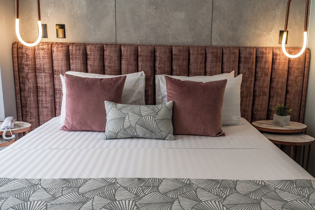 Cama bem arrumada com colcha listrada, duas almofadas grandes e uma almofada estampada. Há duas mesas de cabeceira, uma com um telefone e outra com uma pequena planta. A cabeceira é de madeira com acabamento texturizado.