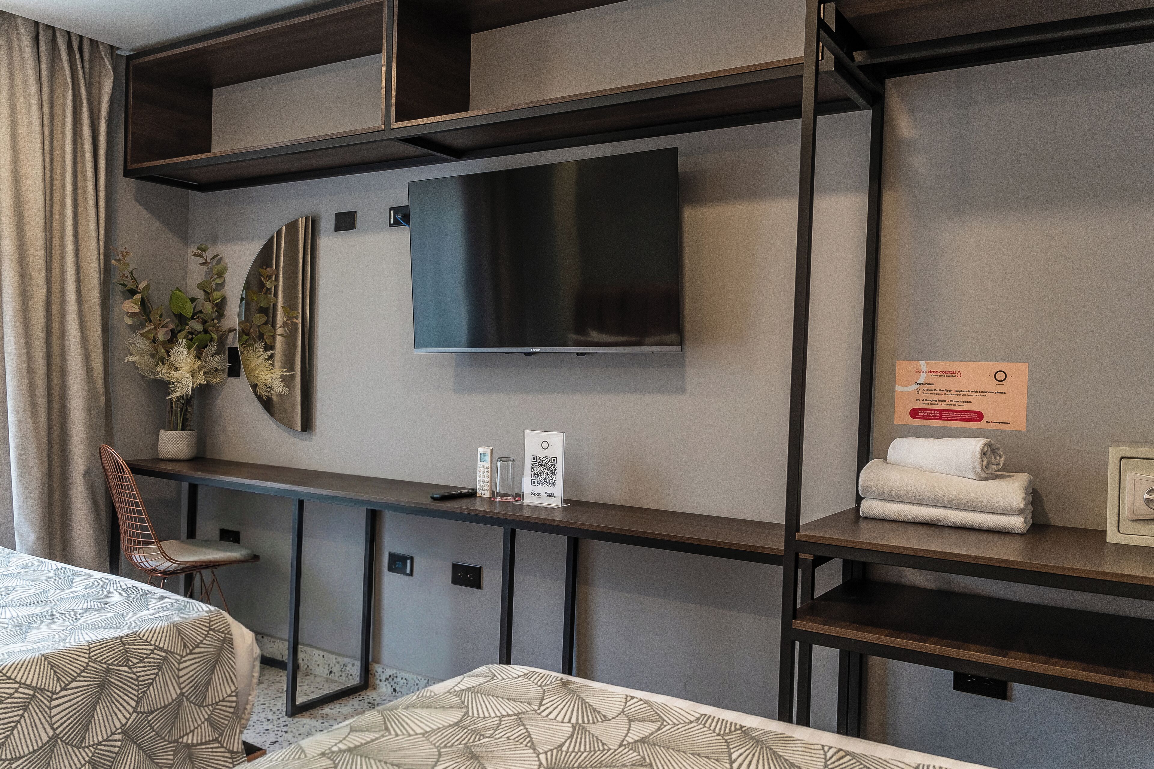 Une chambre d’hôtel moderne dotée d’une grande télévision à écran plat, d’un bureau avec un miroir, d’une chaise et d’un lit avec une literie à motifs.