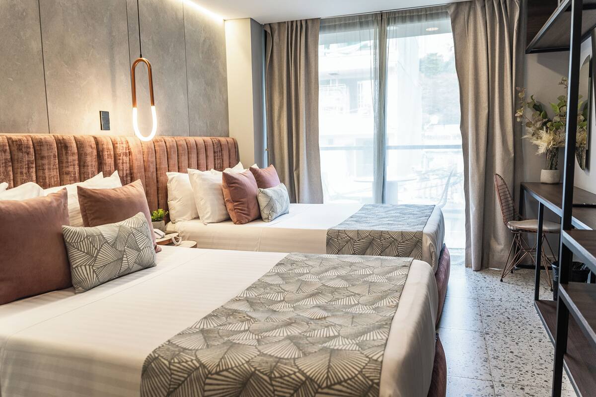 Quarto de hotel moderno com duas camas, uma janela grande com cortinas, uma cabeceira de madeira e uma pequena área de estar com uma cadeira e mesa.