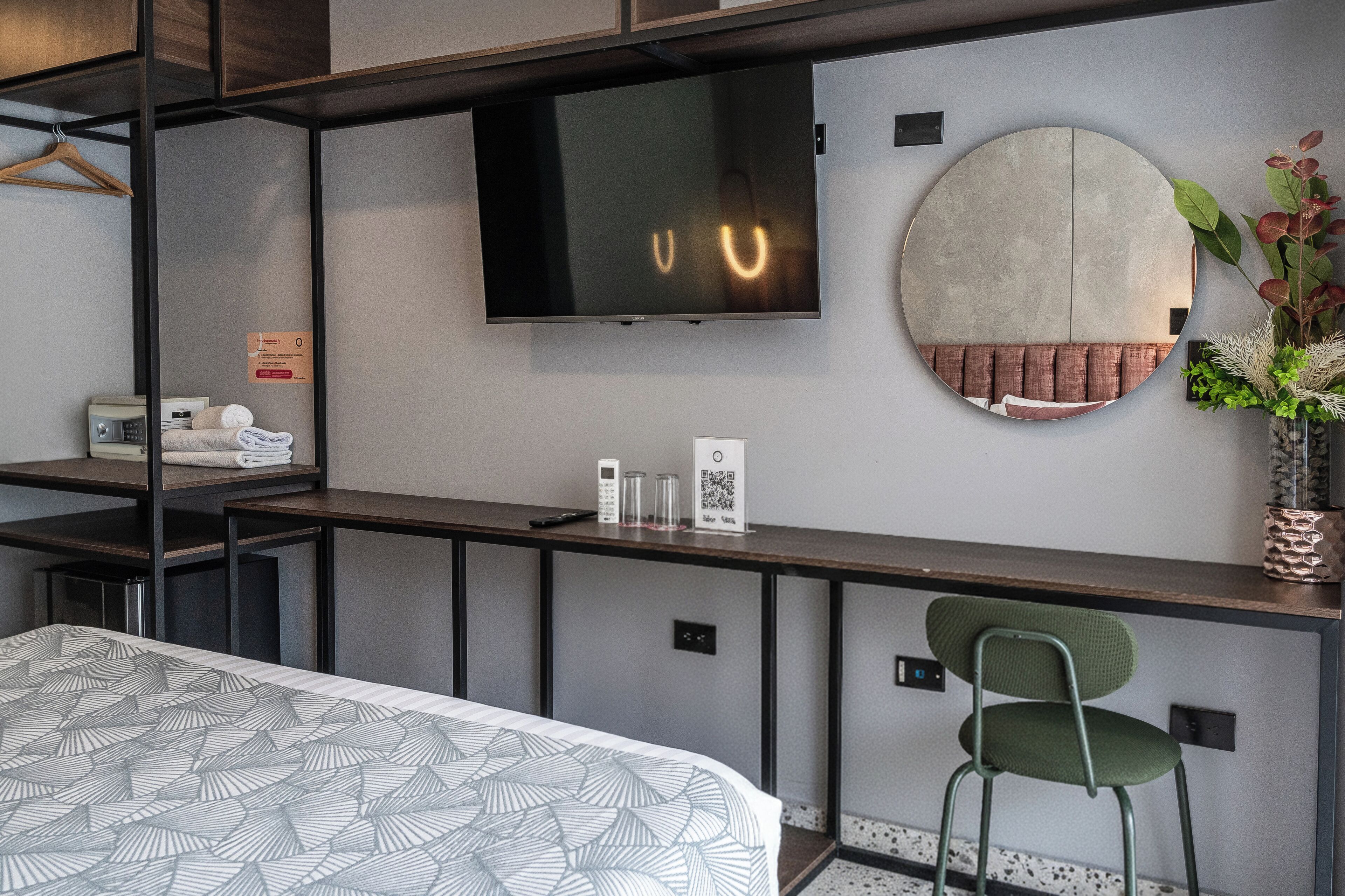 Quarto de hotel moderno com cama, escrivaninha, televisão de tela plana, espelho redondo, cadeira e uma pequena planta.